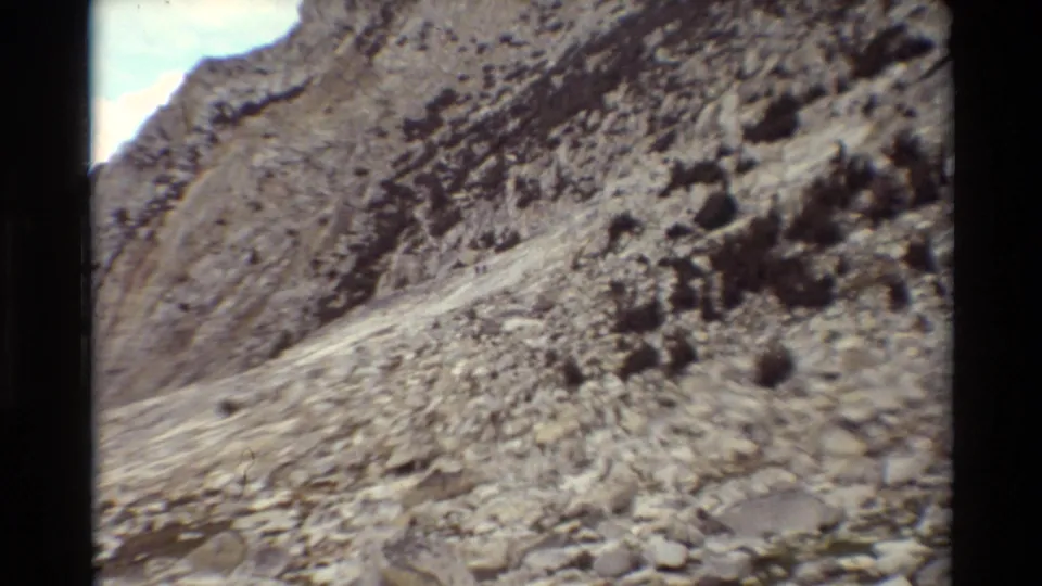 The camera holds on a steep rocky trail across a scree-covered hillside, with scrub and soft 8mm color fading.