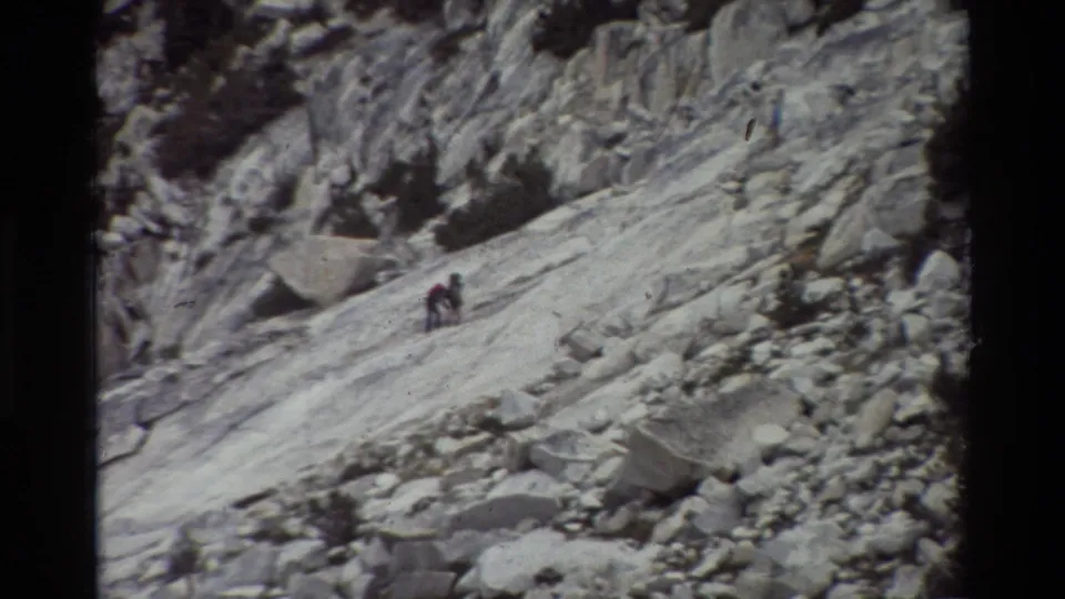Two distant hikers are close together on a steep exposed rock face in a grainy 1981 8mm mountain travel frame.