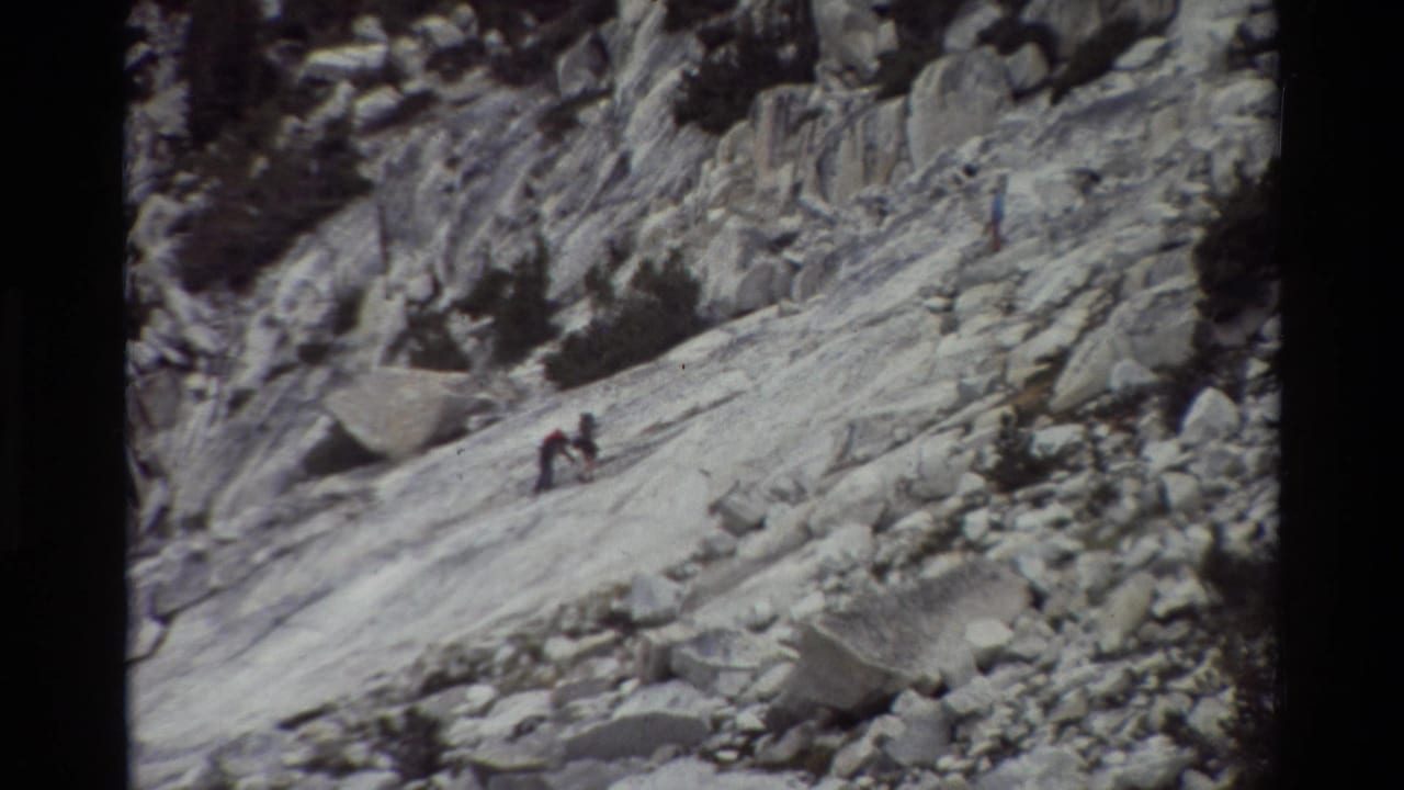 1981 8mm home-movie frame of small hikers moving across a steep rocky mountain slope, with pale stone slabs and scattered low shrubs.