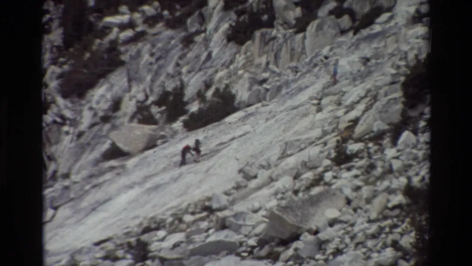 1981 8mm home-movie frame of small hikers moving across a steep rocky mountain slope, with pale stone slabs and scattered low shrubs.