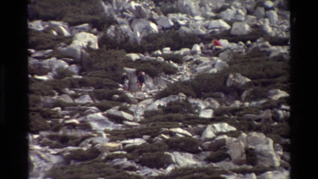 Small hikers moving through boulders and low brush below a rocky ridge.