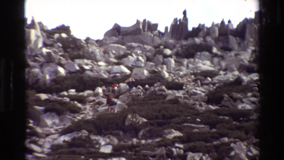 A grainy 8mm frame shows hikers low in the scene crossing a boulder-strewn slope with shrubs and jagged rocks above.