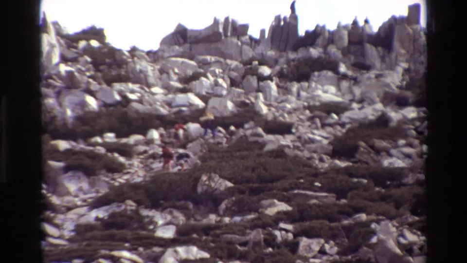 The final 8mm frame shows small hikers in the rocky brush, with large pale boulders, dark shrubs, and jagged ridge shapes behind them.