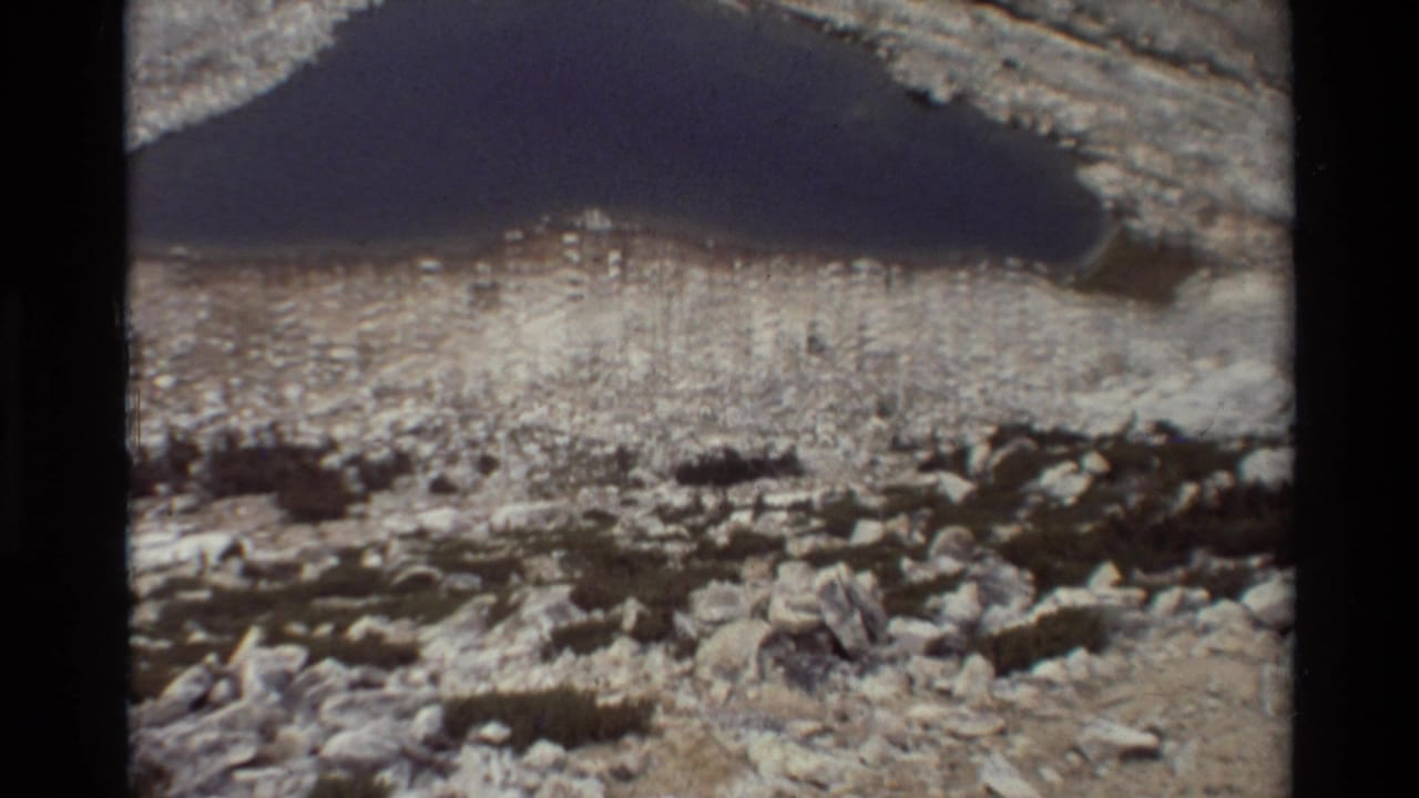 1981 8mm frame of a dark alpine lake set among pale rocky terrain, with boulders and scrub in the foreground.