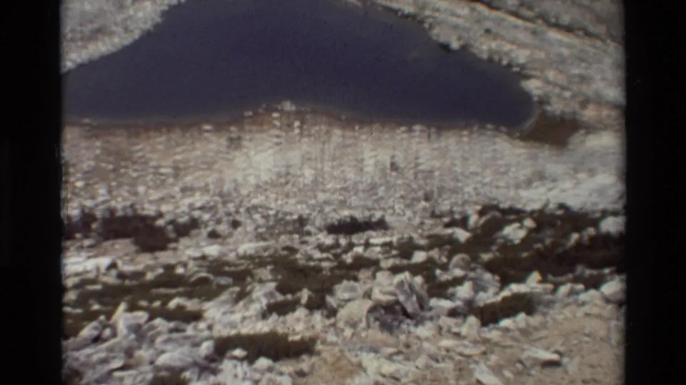 1981 8mm frame of a dark alpine lake set among pale rocky terrain, with boulders and scrub in the foreground.