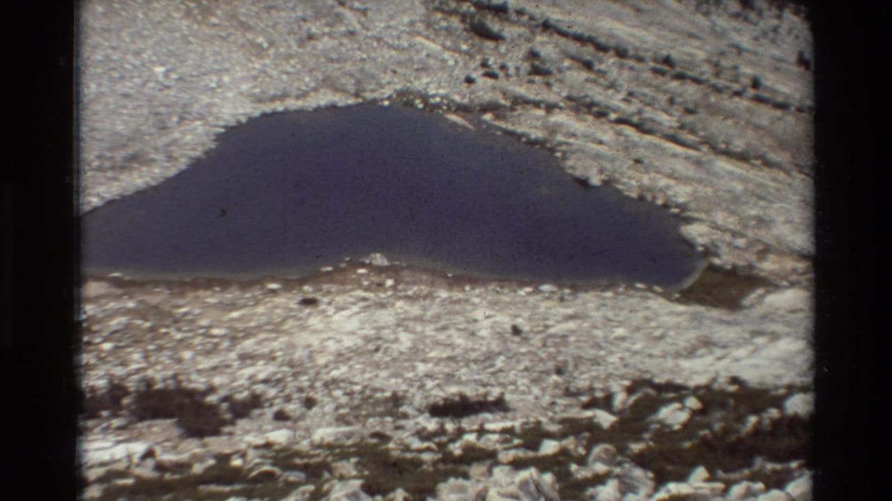 A wider 1981 8mm view of a small dark alpine lake bordered by pale rock and scattered boulders.