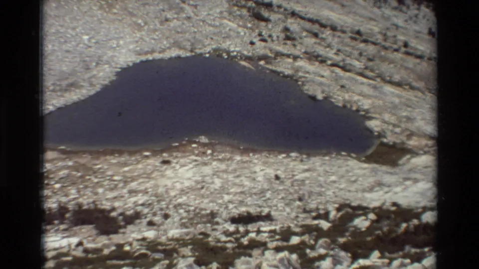 A wider 1981 8mm view of a small dark alpine lake bordered by pale rock and scattered boulders.