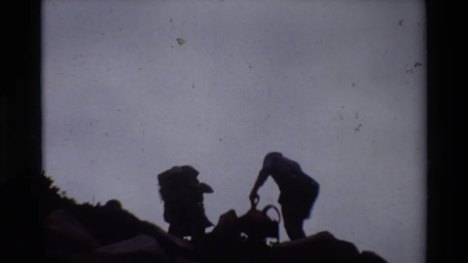Silhouetted hikers in 1981 home-movie footage climb over rocks, their backpacks visible against the bright sky.