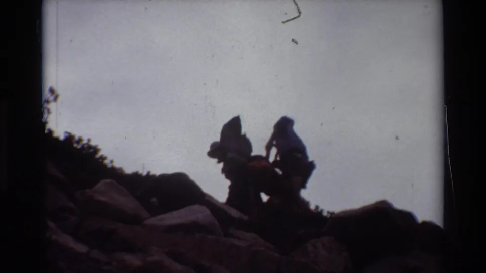 Two silhouetted hikers on a rocky summit area, one appearing to raise an arm while looking out from the ridge.