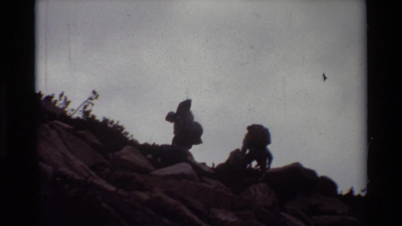 Two hikers with packs continue across uneven rocks under a pale sky.