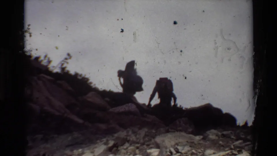 Two backpackers are seen against a washed sky on a rugged slope in this 8mm home-movie frame.