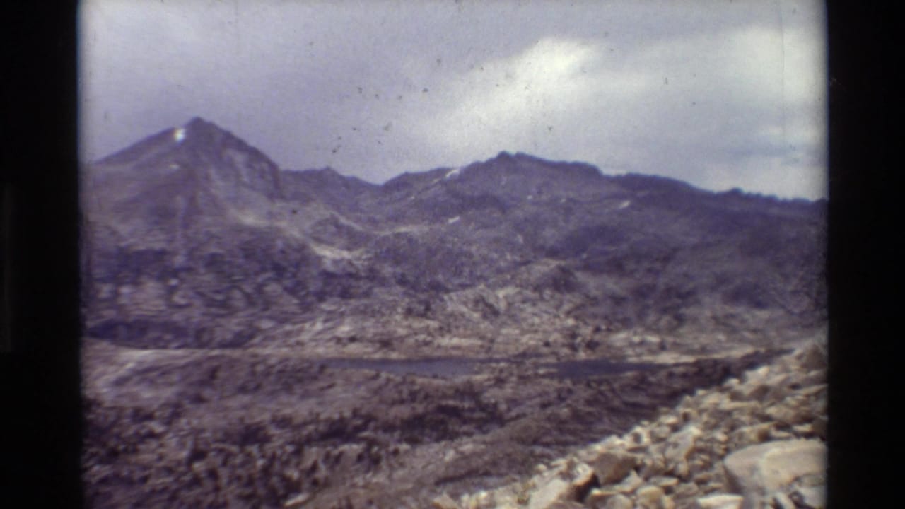1981 8mm home-movie view of rugged mountains and a small alpine lake, with rocky foreground and cloudy sky.