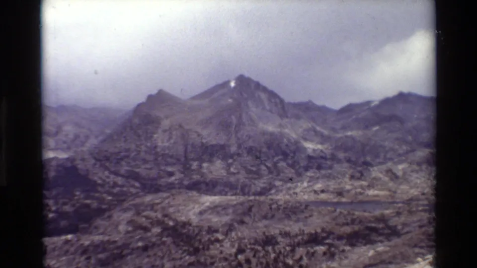 Closer 8mm view of jagged alpine peaks above a small lake, with faded color and soft home-movie focus.