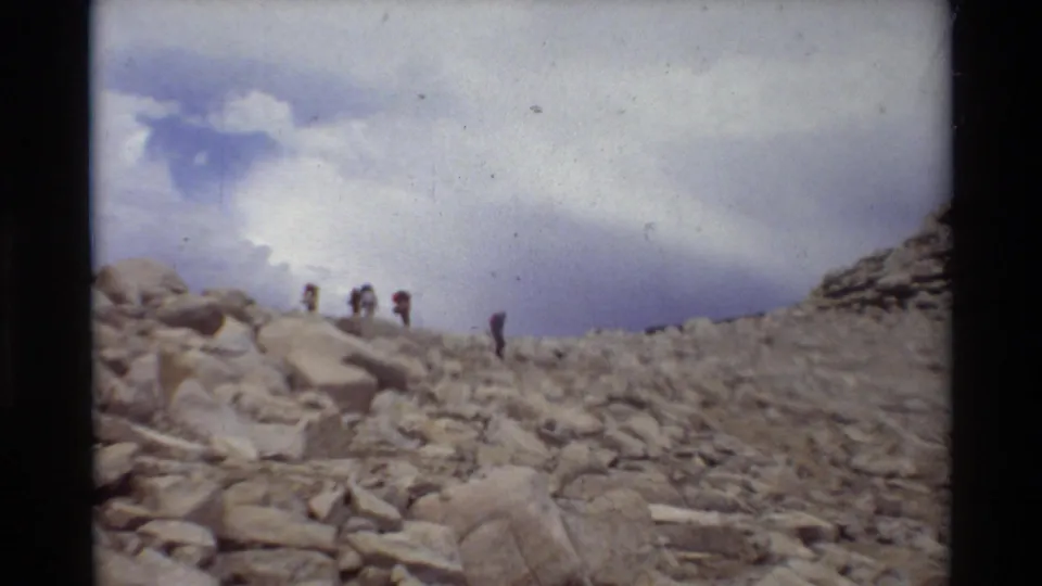Several distant hikers with packs appear to climb a loose rocky slope in the 1981 8mm mountain home movie.