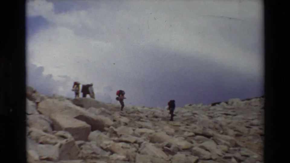 Several hikers carrying packs cross uneven alpine rocks, with the camera looking upward along the slope.