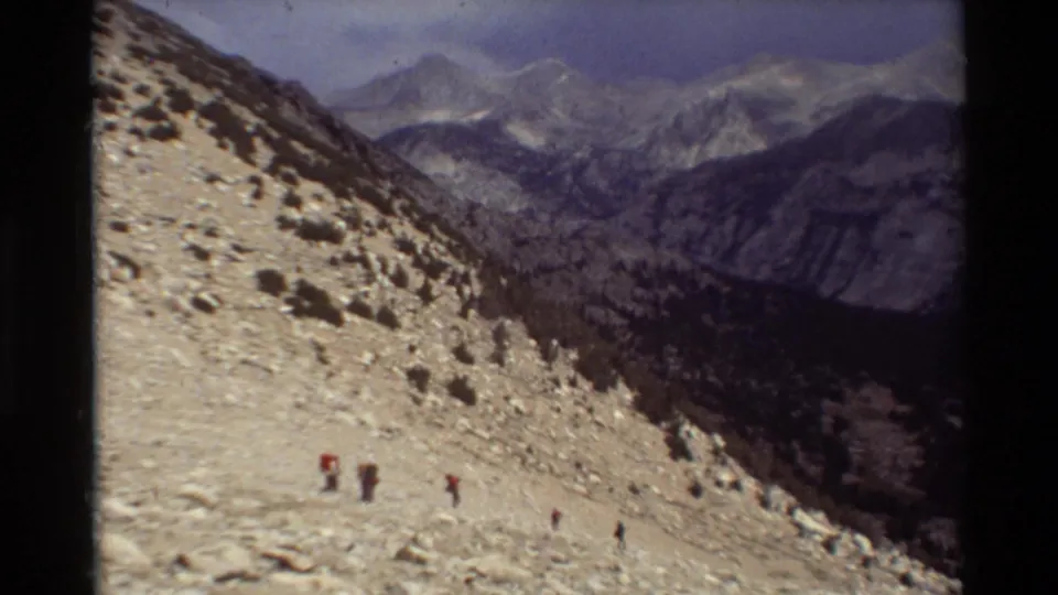 A wide 8mm view shows tiny hikers on a broad rocky mountainside with a valley and distant peaks beyond.