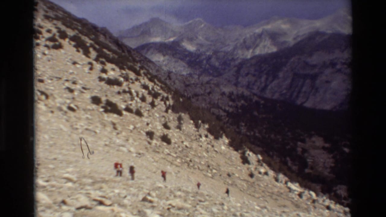 Hikers descend a rocky mountain slope with distant peaks and a valley beyond.
