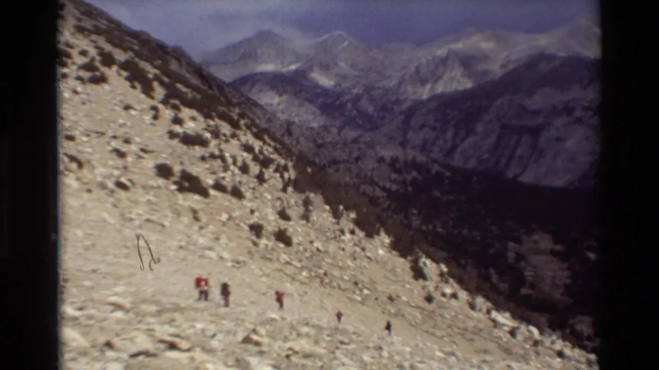 1981 8mm home-movie frame of hikers moving down a pale rocky alpine slope above a deep mountain valley.