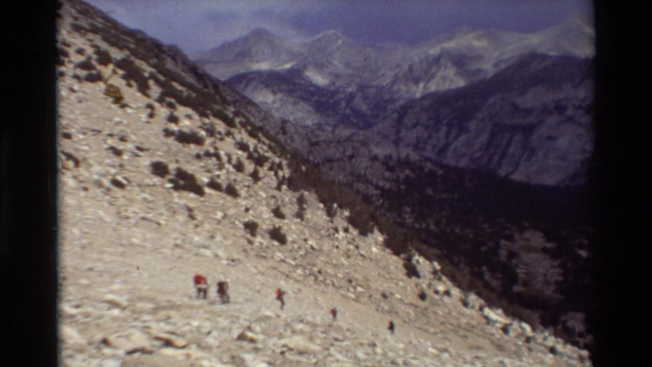 Hikers appear as small figures on a sunlit rocky slope, with layered mountain ridges behind them.