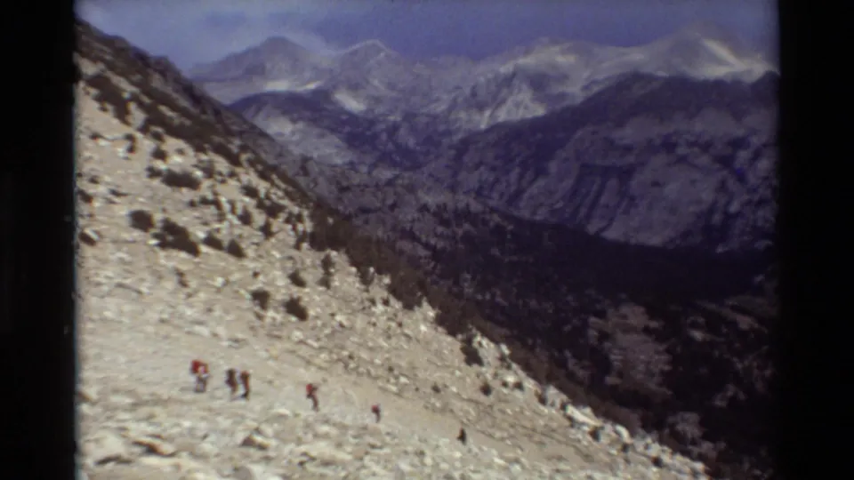 The home-movie view widens across hikers descending uneven alpine ground above a dark valley and distant mountains.