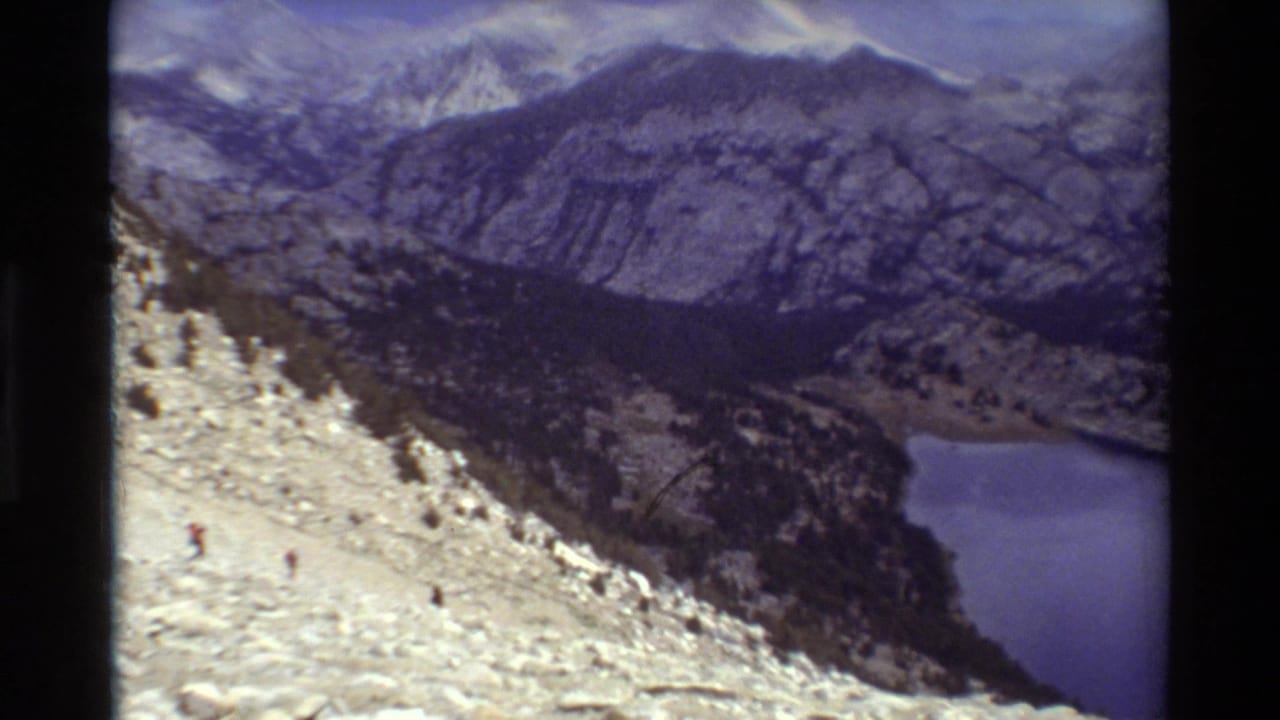 Rocky alpine slope with tiny hikers above a blue lake and rugged mountains.