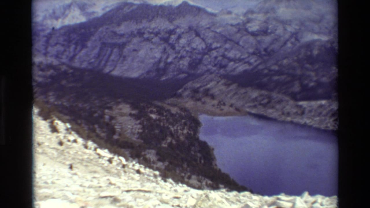 High 8mm travel-film viewpoint across a blue-purple alpine lake, pale rocks, a dark valley edge, and rugged mountain ridges.