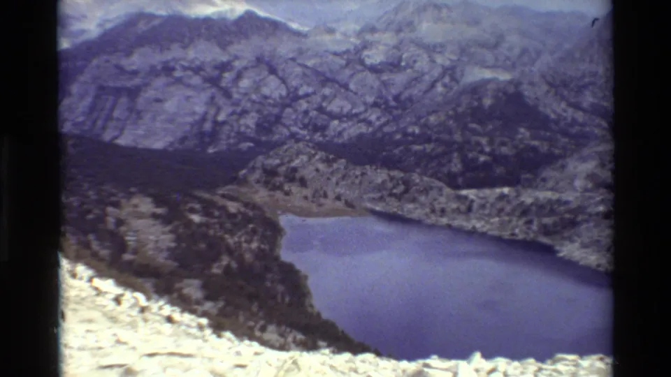 Soft, grainy 8mm frame showing an alpine lake below pale rocky foreground, dark valley forms, and hazy mountain ridges.