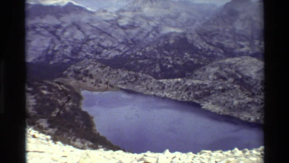 Blue-toned 8mm home-movie frame of a lake curving through a rocky alpine valley, with rugged peaks and soft exposure shift.