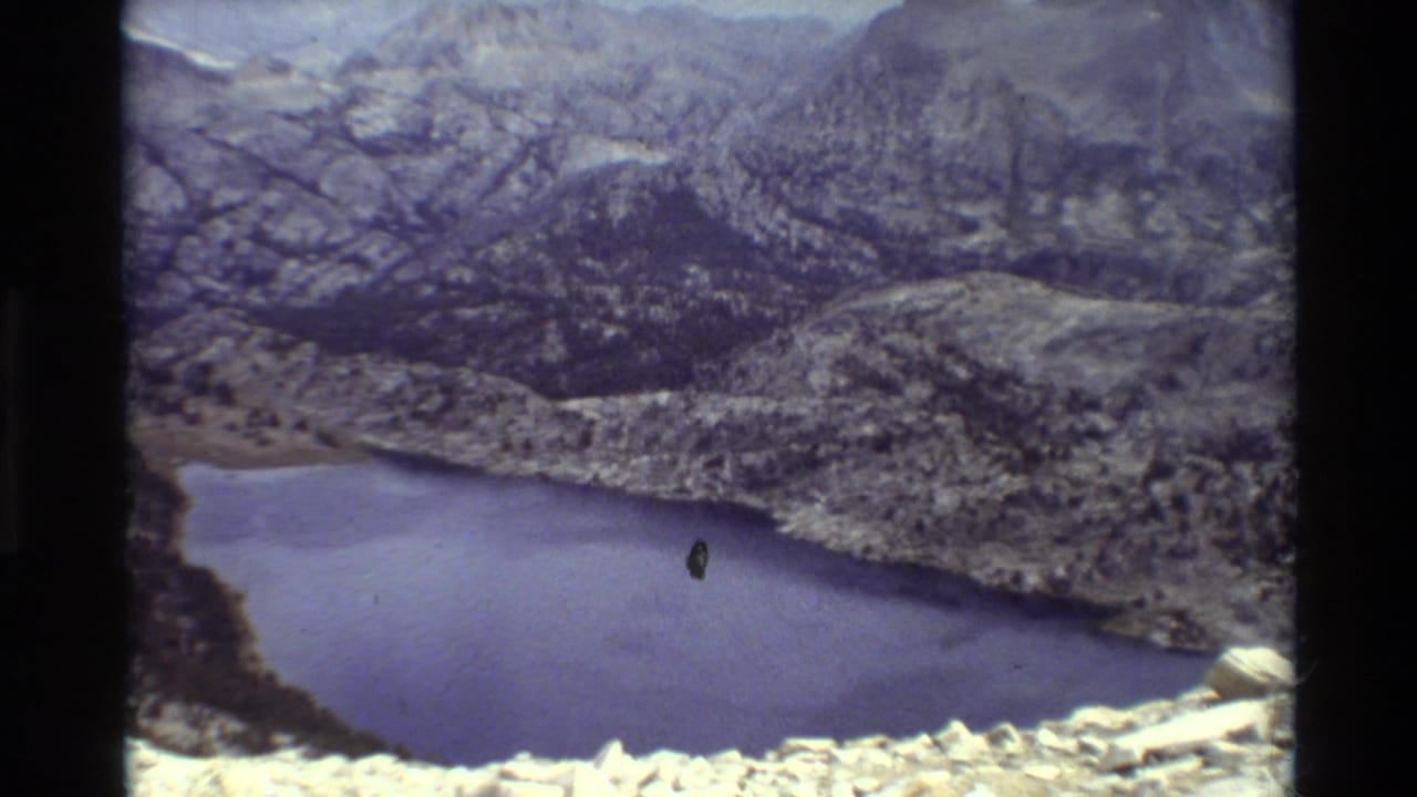 1981 8mm frame showing a blue mountain lake below rugged rock terrain, with muted color and visible film edge marks.