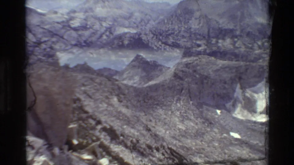 1981 8mm frame looking across a rocky alpine basin toward distant water, sharp ridges, and small snow patches.