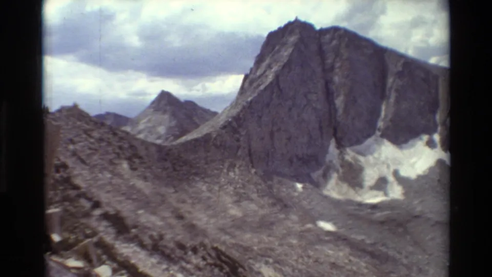 1981 8mm frame of a steep rocky peak under a cloudy sky, with snow patches and barren alpine terrain below.
