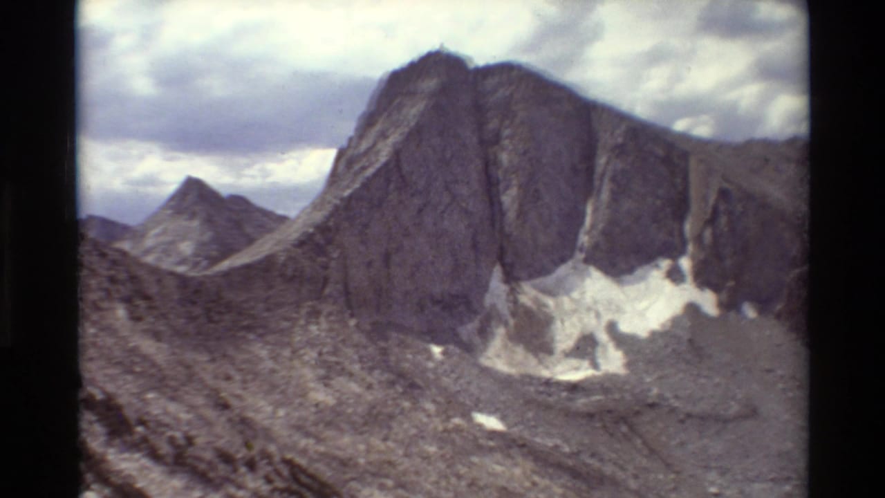 1981 8mm-style mountain view showing steep grey rock faces, talus slopes, small snow patches, and cumulus clouds.