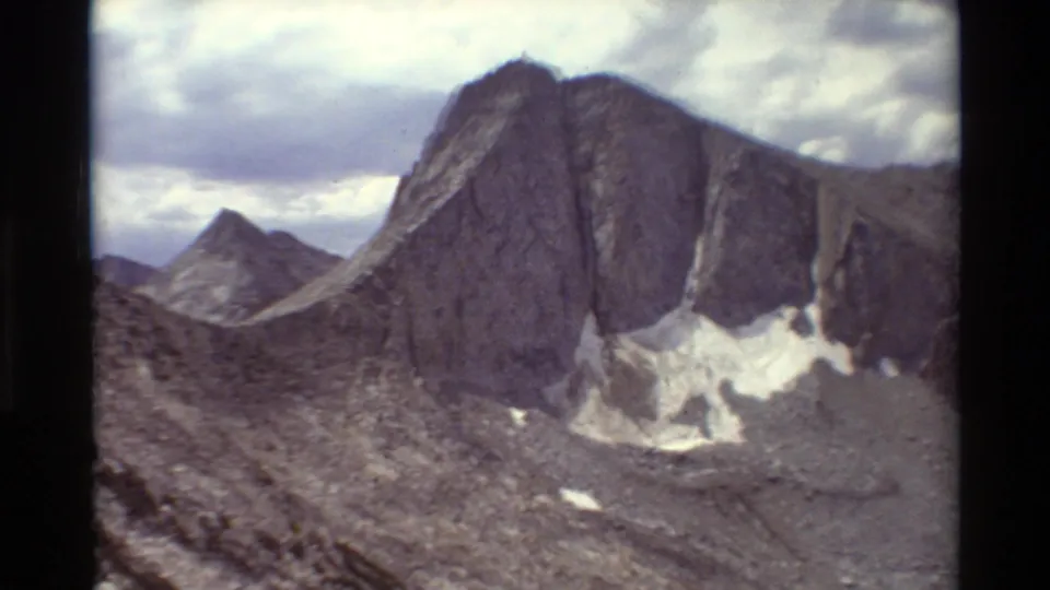 1981 8mm-style mountain view showing steep grey rock faces, talus slopes, small snow patches, and cumulus clouds.