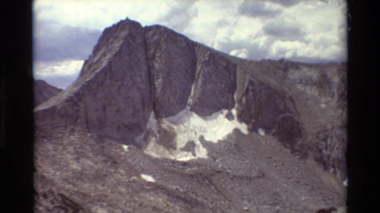 Archival 8mm frame of a steep grey alpine ridge with lingering snow patches and a pale, clouded sky.