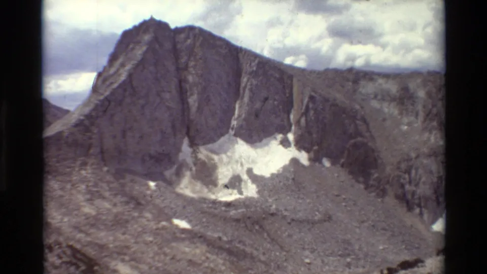 1981 home-movie-style alpine view of a grey mountain wall, rocky slopes, and bright snow patches below clouds.