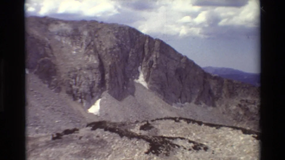 Archival 8mm frame showing a sloping grey ridge, small snowfields, rocky foreground, and distant horizon.