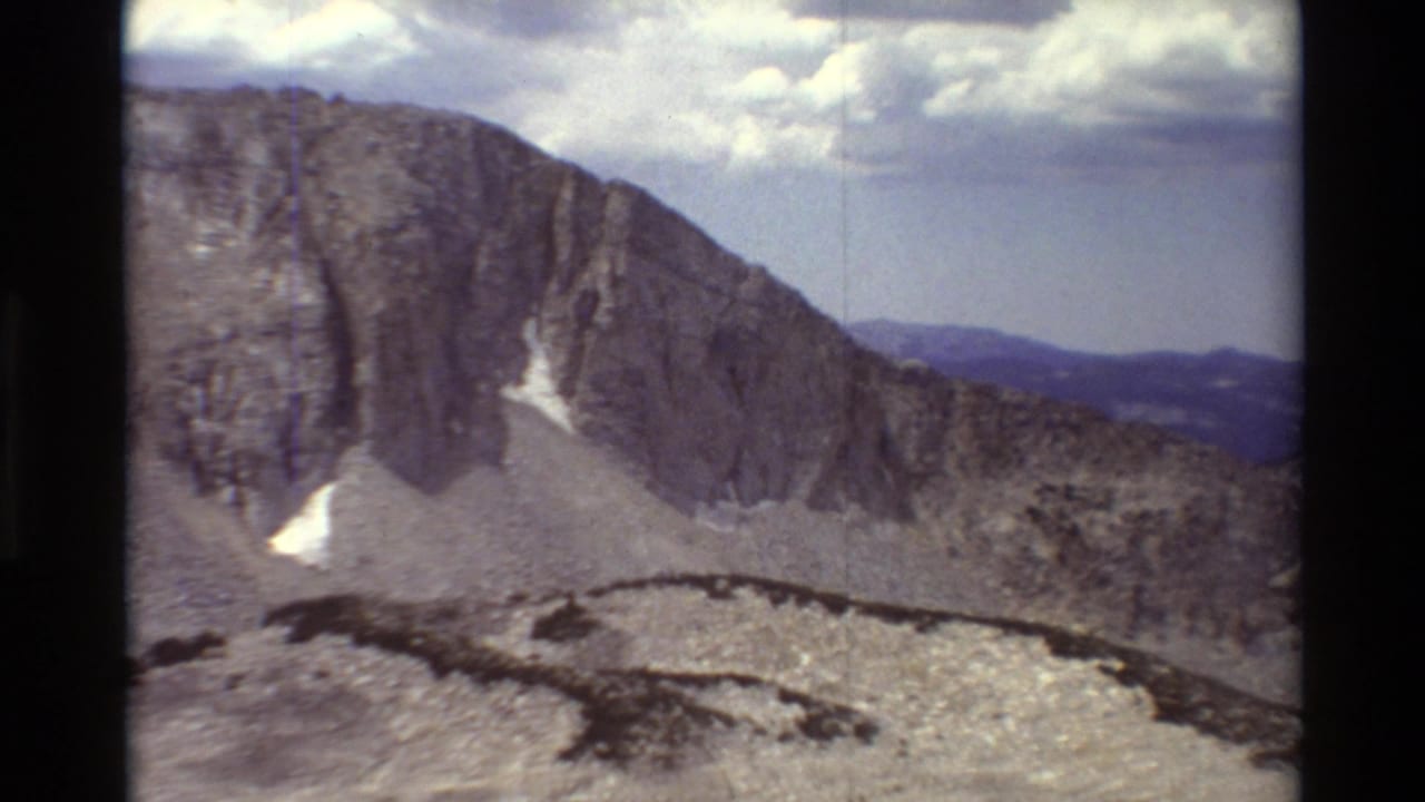 1981 8mm frame of a rugged alpine ridge with cliff faces, pale rock slopes, small snow patches, and dark film edges.