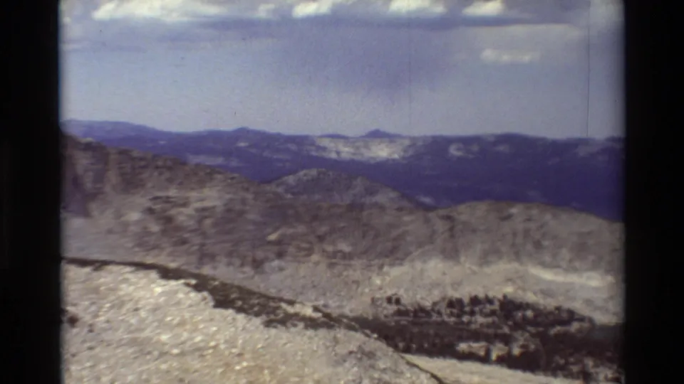 1981 8mm mountain pan frame with a rocky foreground, dark tree line, and distant blue ridges under a pale sky.
