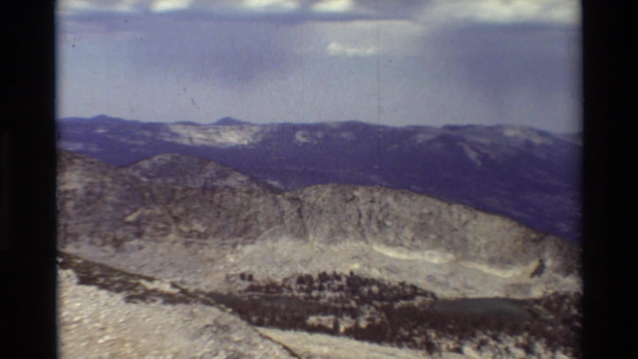 1981 8mm frame of a rocky mountain ridge with layered distant peaks and scattered evergreens, based on supplied British Columbia metadata.