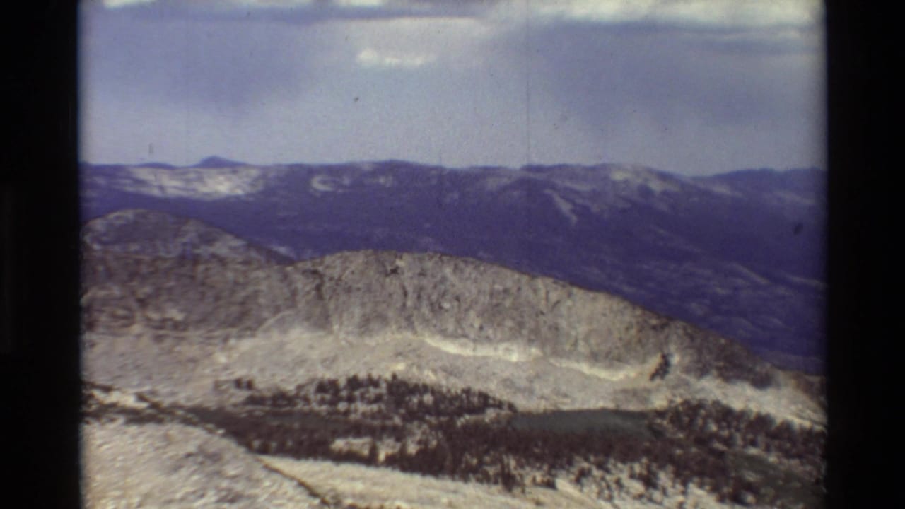 Soft 8mm view of a rugged ridge, dark distant mountains, pale rocky slopes, and sparse evergreen forest.