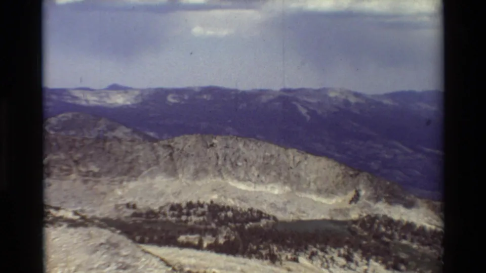 Soft 8mm view of a rugged ridge, dark distant mountains, pale rocky slopes, and sparse evergreen forest.