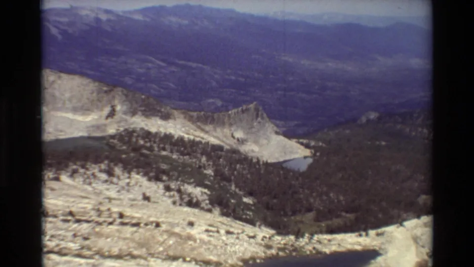 1981 home-movie frame showing a forested mountain valley, rocky slopes, distant ridges, and a small alpine lake below.