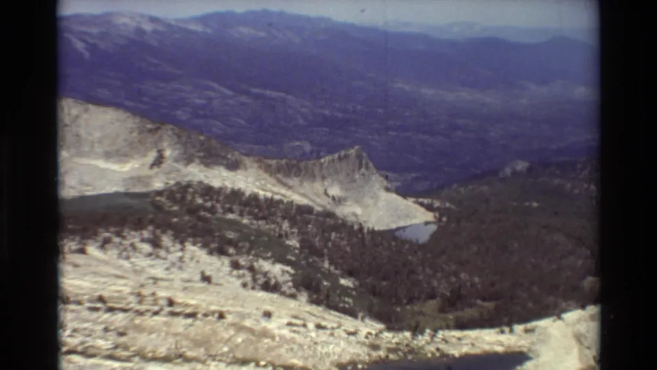 Wide 8mm mountain panorama with a rocky ridge in front, forested valley below, a small lake, and hazy distant peaks.