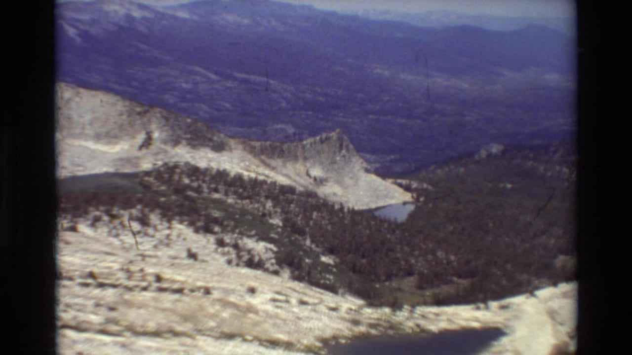 1981 8mm view of a rugged mountain overlook, with snow patches, a forested basin, distant ridges, and a small lake below.