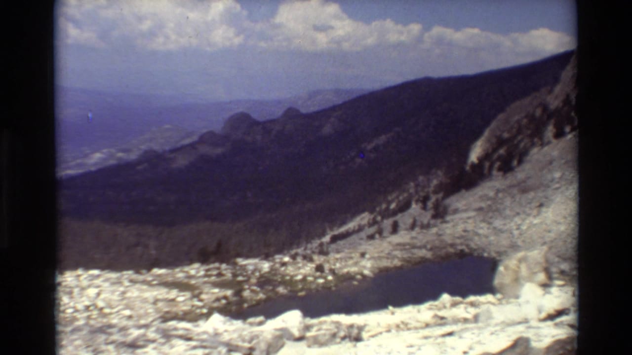 1981 8mm mountain panorama with a rocky snowy foreground, dark alpine lake, forested slopes, and hazy ridges under clouds.