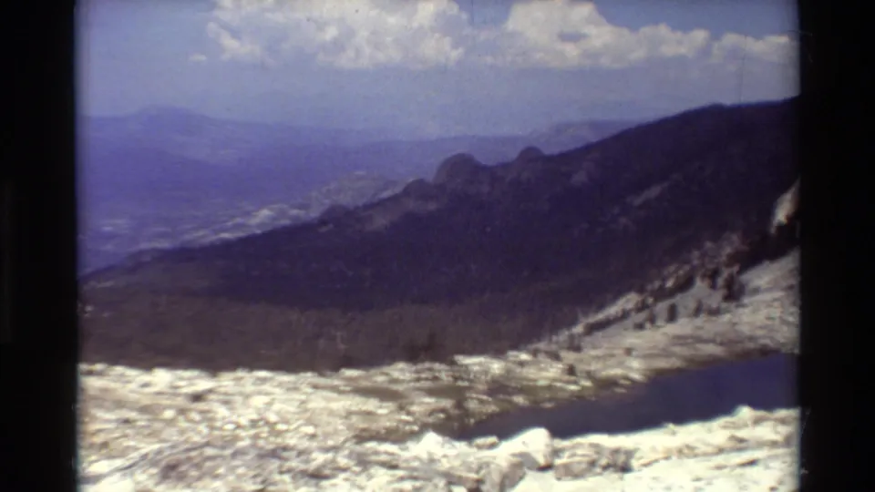 Soft 1981 8mm frame of a mountain valley, showing snow-streaked rock, a dark lake edge, forested slopes, and distant hazy ridges.