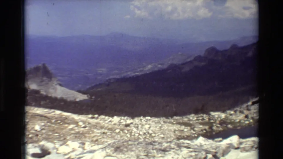 1981 8mm wide view across rocky snow patches toward a forested basin and layered mountain ridges beneath a clouded sky.