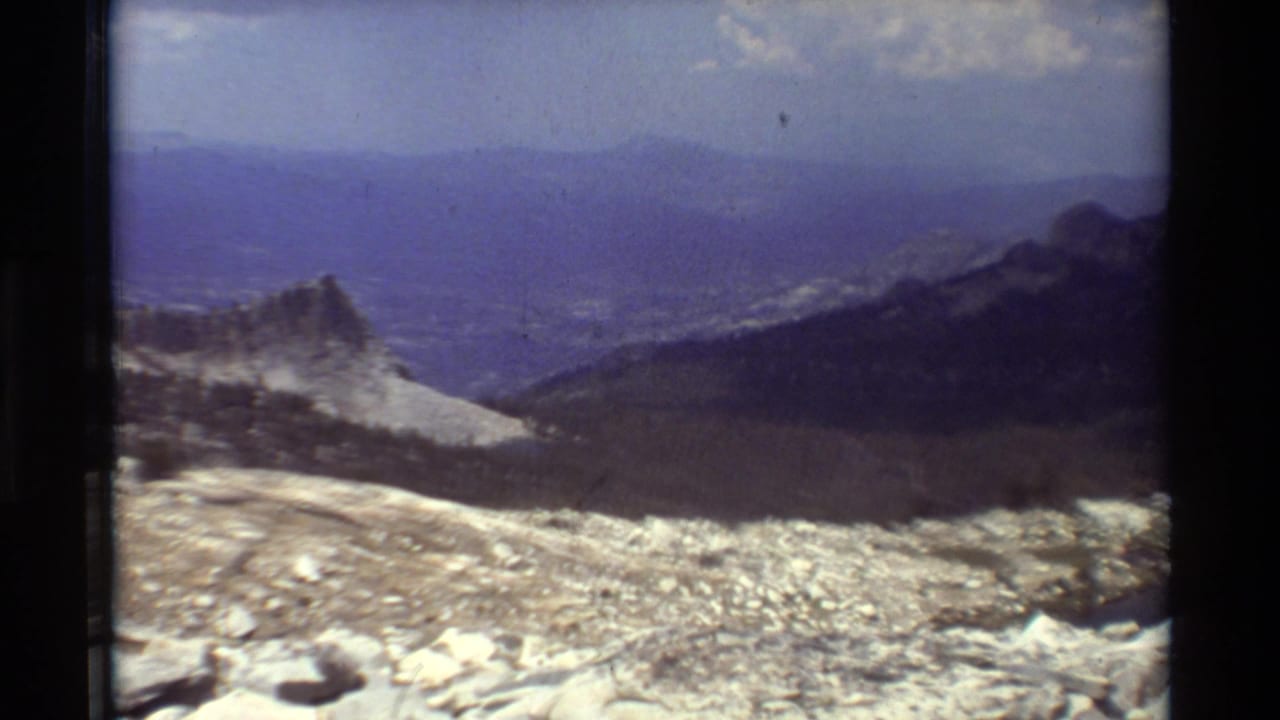 1981 8mm home-movie frame of a rocky high overlook facing forested slopes, a distant valley, blue ridges, and scattered clouds.