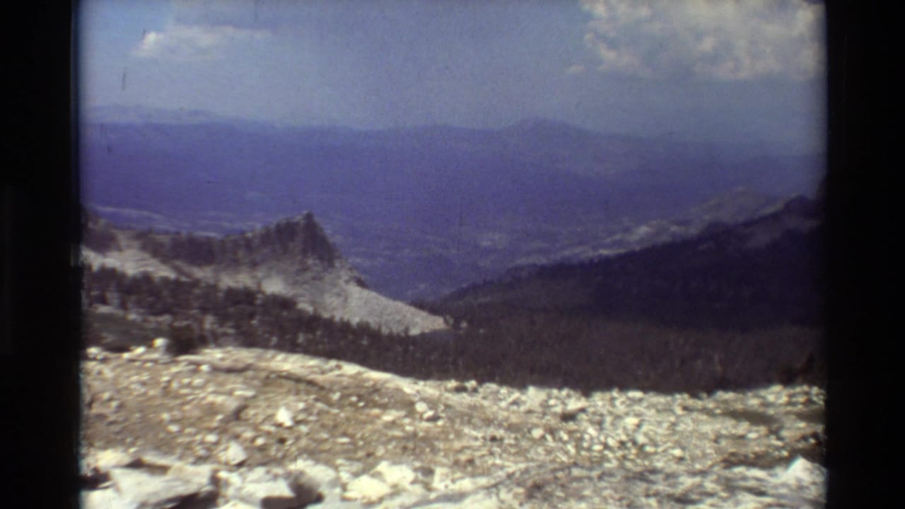 Wide mountain vista with rocks, a ridge, forested slopes, and blue sky.