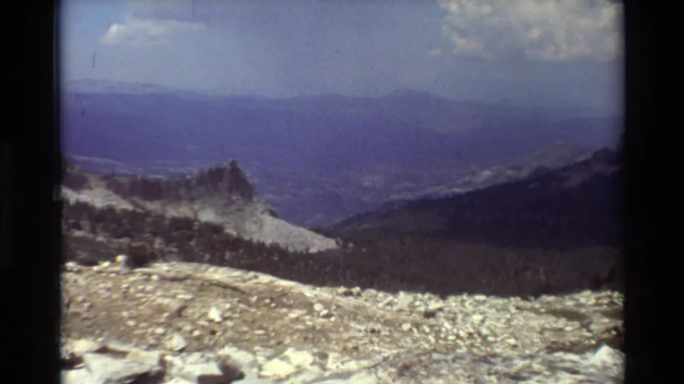 Handheld 8mm view across a stony alpine slope toward a jagged ridge, dark forest below, distant hills, and a bright clouded sky.